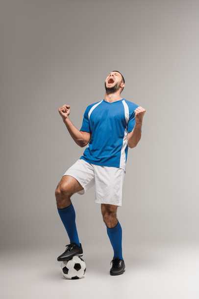 Deportista excitado en ropa deportiva mostrando sí gesto cerca del fútbol sobre fondo gris  - Foto, imagen