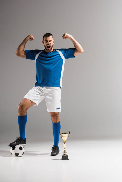 Esportista animado mostrando sim gesto perto de futebol e dourado campeões copo no fundo cinza  - Foto, Imagem