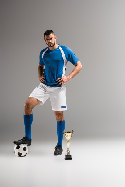 Deportista con las manos en las caderas mirando a la cámara cerca del fútbol y la copa de campeones sobre fondo gris  - Foto, imagen