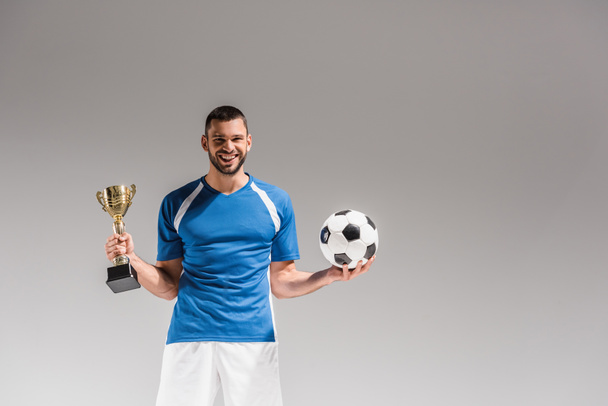 Cheerful sportsman holding football and golden champions cup on grey background  - Photo, Image