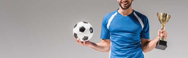 Vista recortada del deportista sonriente sosteniendo la copa de campeones y el fútbol aislado en gris, bandera  - Foto, Imagen
