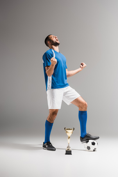 Sportsman celebrating triumph near football and champions cup on grey background  - Fotografie, Obrázek