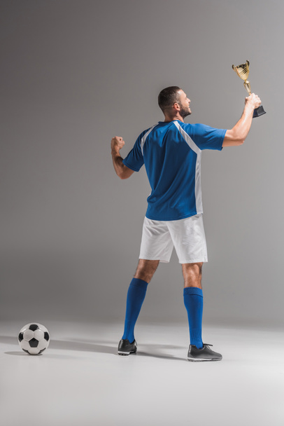 Visão traseira do esportista sorridente segurando troféu dourado perto de futebol no fundo cinza  - Foto, Imagem