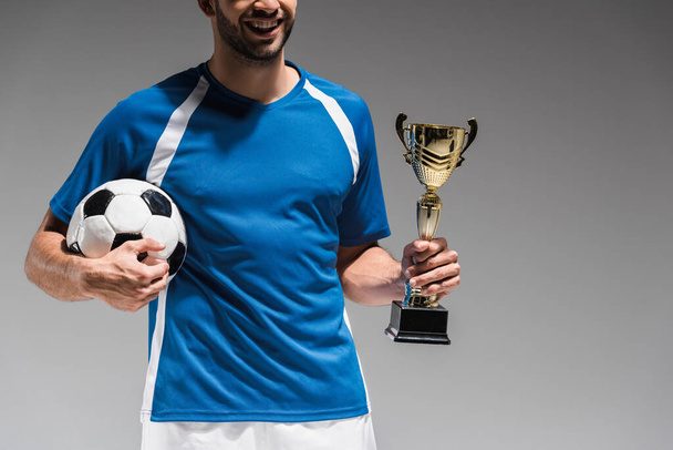 Oříznutý pohled na usmívajícího se sportovce s pohárem šampionů a fotbalem izolované na šedé - Fotografie, Obrázek