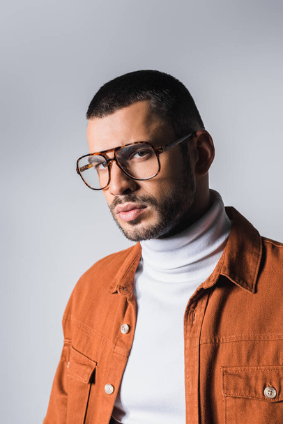 Trendy man in eyeglasses and terracotta jacket looking at camera isolated on grey - Foto, imagen