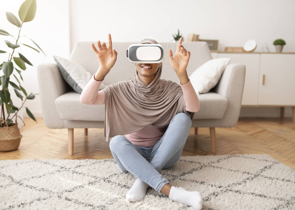 Weekend pastimes. Young Muslim woman in hijab interacting with virtual reality, wearing VR headset at home - Photo, Image