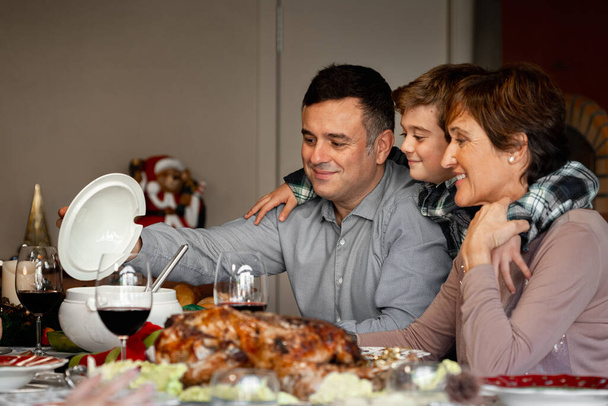 Portret van zoon, kleinzoon en grootmoeder zittend aan de kerstThanksgiving tafel, kijkend naar het heerlijke eten. Soep, kalkoen en rode wijn. Samen zijn, kerstvakantie vieren.. - Foto, afbeelding