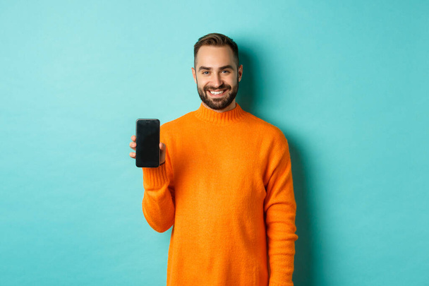 Chico barbudo guapo en suéter naranja, mostrando la pantalla del teléfono inteligente y sonriendo, mostrando promo en línea, fondo turquesa - Foto, imagen