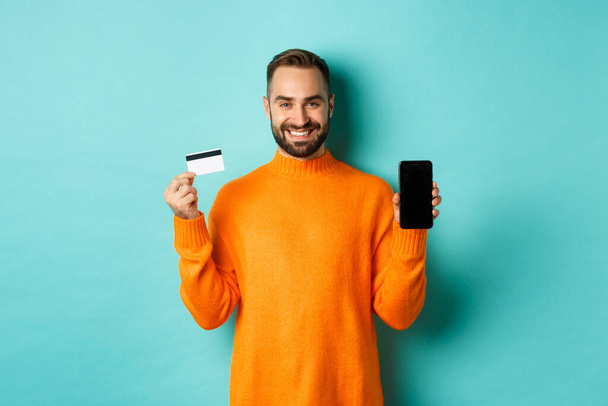Online shopping. Happy attractive guy showing mobile phone screen and credit card, smiling satisfied, standing over light blue background - Valokuva, kuva