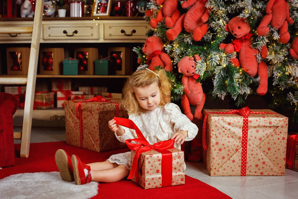 A little blonde girl in a white dress opens a gift sitting near the Christmas tree and lights of garlands. - Foto, Imagen