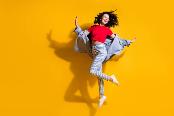 Full length photo of cute lady look empty space jump wear striped jeans cropped red t-shirt jacket sneakers isolated yellow color background - 写真・画像