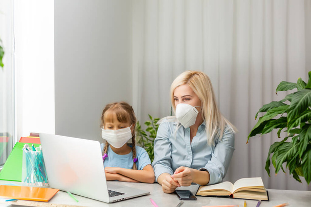 Jonge vrouw met medisch masker wijzend naar gegevens op laptop voor klein meisje terwijl ze aan tafel zit en samen aan huiswerk werkt - Foto, afbeelding