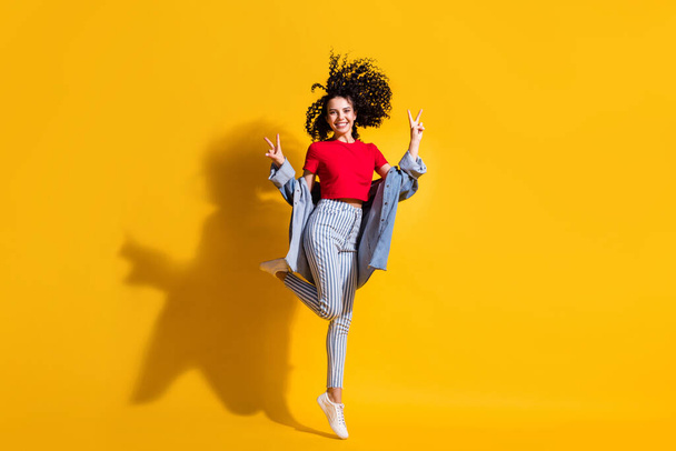 Full length photo of girl stay tiptoe show v-sign wear striped jeans cropped red t-shirt jacket sneakers isolated yellow color background - Фото, зображення