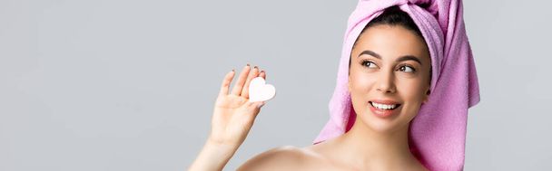 happy beautiful woman with towel on hair holding heart shaped sponge isolated on grey, banner - Photo, Image