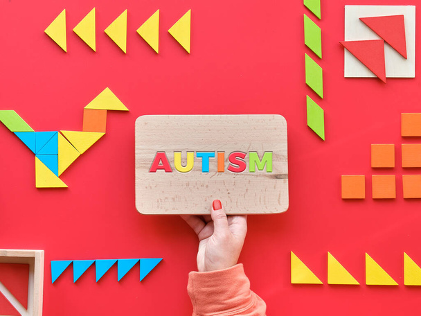 Creative design for Autism World Day on April 2, text on lightbox. Tangram elements scattered and arranged in pictogram on red background. Hand hold wooden board with text Autism. - Foto, Bild