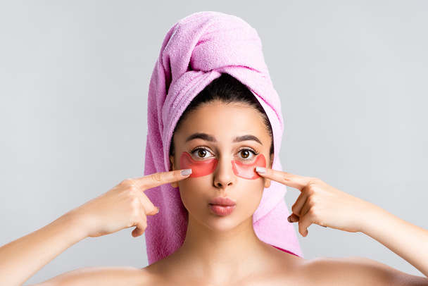 mooie vrouw met handdoek op haar wijzend naar hydrogel ooglapjes op gezicht geïsoleerd op grijs - Foto, afbeelding
