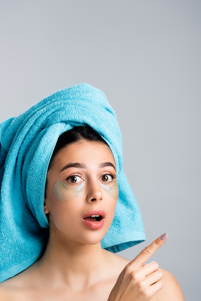 impactado hermosa mujer con toalla azul en el cabello y parches de hidrogel en la cara señalando a un lado aislado en gris - Foto, Imagen