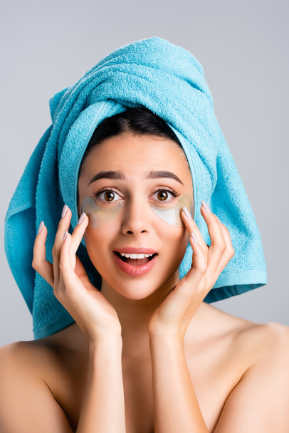 užaslá krásná žena s modrým ručníkem na vlasech a hydrogel oční náplasti na tváři izolované na šedé - Fotografie, Obrázek