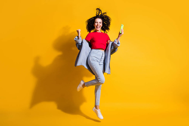 Photo of lady hold smartphone open mouth raise fist jump wear striped jeans red t-shirt jacket sneakers isolated yellow color background - Foto, afbeelding