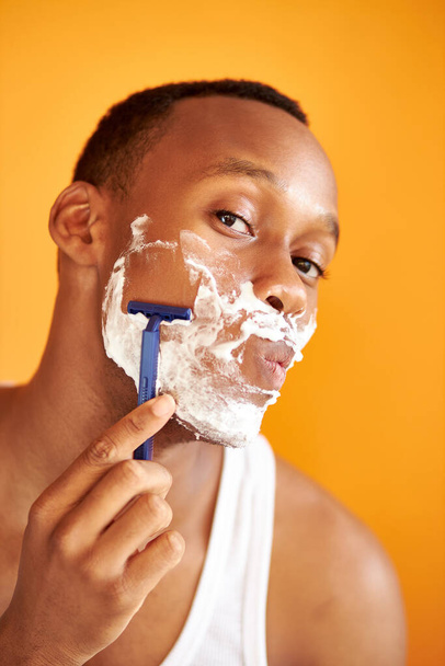 Porträt eines afrikanischen Mannes, der sein Gesicht mit Schaum und Rasiermesser isoliert auf orangefarbenem Hintergrund rasiert - Foto, Bild