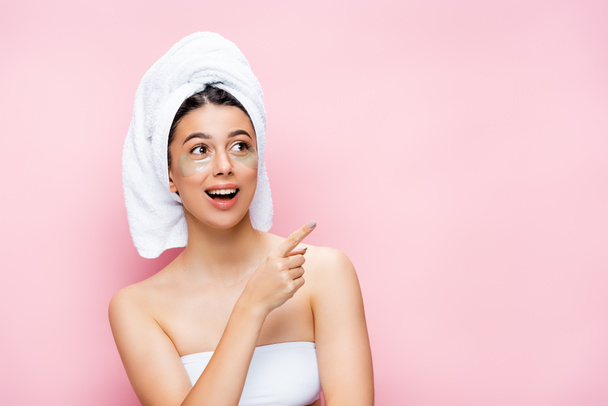 excited beautiful woman with towel on hair and hydrogel eye patches on face pointing aside isolated on pink - 写真・画像
