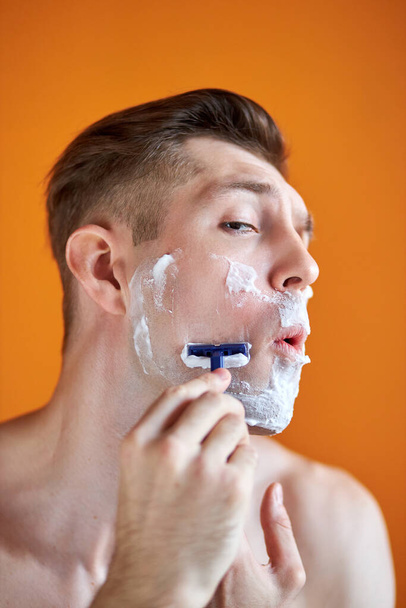 portrait of caucasian male shaving his face with foam and razor isolated on orange background - Photo, Image