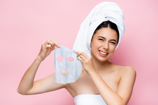 disgusted beautiful woman with towel on head holding mask sheet isolated on pink - Photo, Image