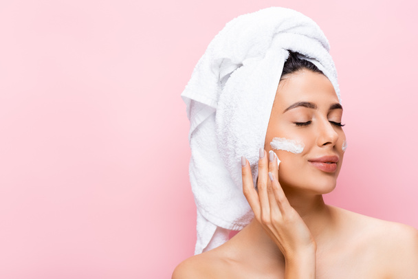 krásná žena s ručníkem na vlasech, zavřené oči a kosmetický krém na tváři izolované na růžové - Fotografie, Obrázek