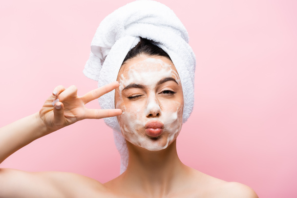 beautiful woman with towel on hair and foam on face grimacing isolated on pink - Photo, Image