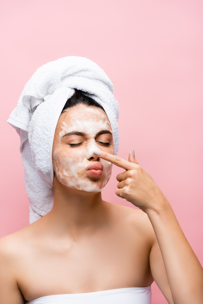 krásná žena s ručníkem na vlasy a pěna na tváři dotýká nosu izolované na růžové - Fotografie, Obrázek