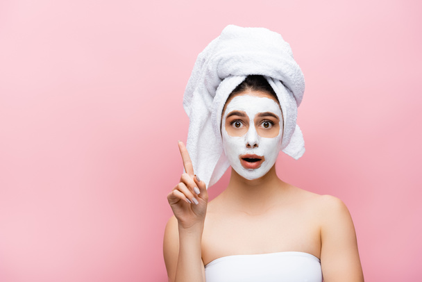 shocked beautiful woman with towel on head and clay mask on face pointing aside isolated on pink - Фото, зображення