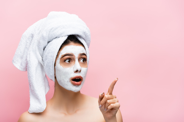 šokovaná krásná žena s ručníkem na hlavě a hliněné masky na tváři ukazuje stranou izolované na růžové - Fotografie, Obrázek