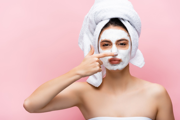 beautiful woman with towel on head and clay mask on face touching nose isolated on pink - Фото, изображение