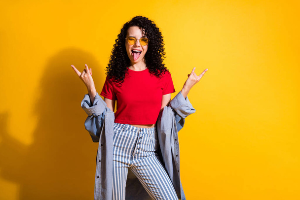 Photo of pretty lady blink eye tongue-out flirty show horns wear red t-shirt striped jeans jacket isolated yellow color background - Fotografie, Obrázek