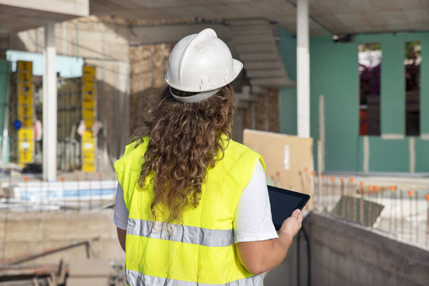 Rear view of woman architect wearing neon safety vest and white helmet and holding an electronic device while inspecting the quality and checking out the progress of the construction in front of her - Фото, зображення