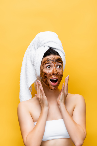 shocked beautiful woman with towel on head and coffee mask on face isolated on yellow - Фото, изображение