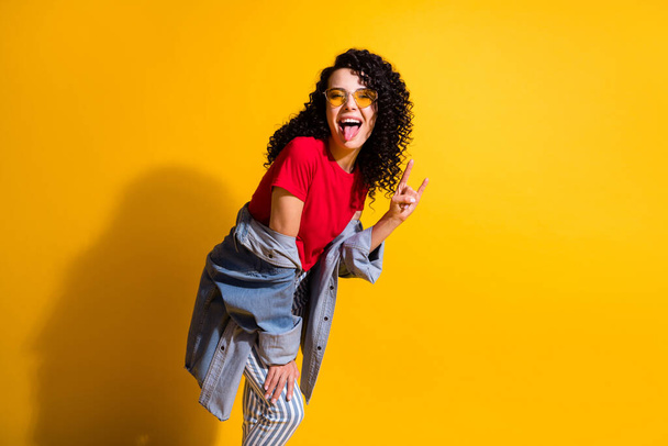 Photo of lovely young lady tongue-out show horns wear red t-shirt striped jeans jacket sunglass isolated yellow color background - Foto, afbeelding