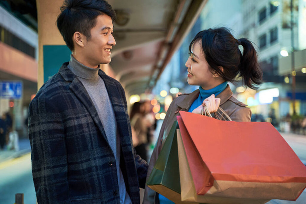 szczęśliwa i kochająca młoda azjatycka para stojąca na dworcu autobusowym z torbami na zakupy w ręku - Zdjęcie, obraz