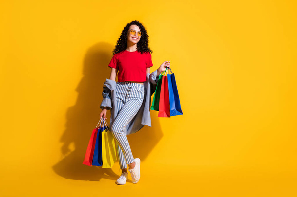 Photo of lady look up empty space hold shop packets wear red t-shirt striped jeans jacket sunglass sneakers isolated yellow color background - Fotó, kép