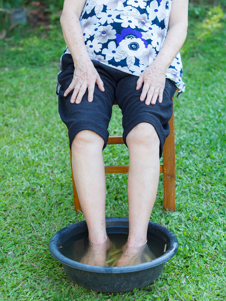 Vanhempi nainen on nilkkakipu hoito kylpylä jalka yrttejä puutarhassa. Lihakset liottamalla lämmintä vettä, joka on keitetty yrttejä. Ikääntyneiden käsite ja terveydenhuolto. - Valokuva, kuva