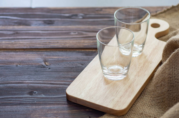 Side view of two empty glasses on small wooden cutting board placed near brown sackcloth on brown wooden table. Glasses cup for drinks. Beverage concept. - Fotoğraf, Görsel