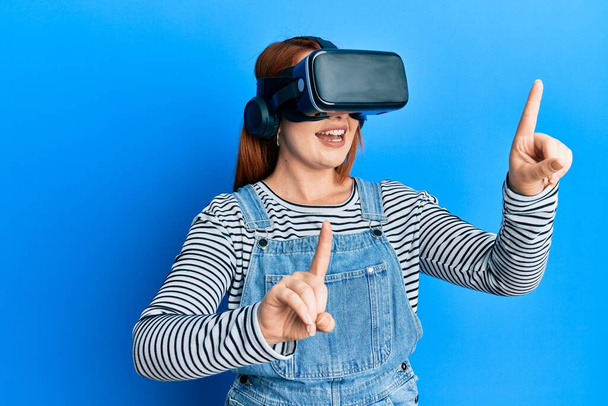 Mujer pelirroja joven usando gafas virtuales 3d de pie sobre fondo azul aislado. - Foto, imagen