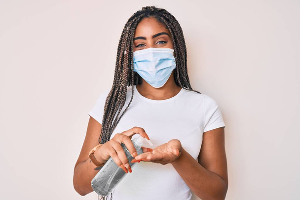 Молода афро-американська жінка з плечима в медичній масці за допомогою ручного санітиста посміхається з щасливою і прохолодною посмішкою на обличчі. з зубами.  - Фото, зображення