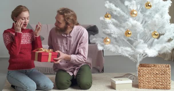Famiglia caucasica che festeggia il nuovo anno insieme a casa, giovane donna bionda seduta a coprirsi gli occhi con le mani uomo barbuto caucasico che dà alla moglie fidanzata regalo di Natale scatola regalo rossa - Filmati, video