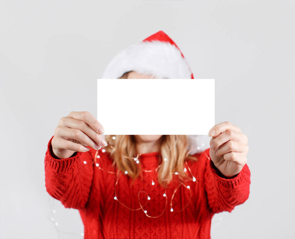 Noel Baba şapkalı genç kadın yüzünü beyaz kağıtla örtüyor. - Fotoğraf, Görsel