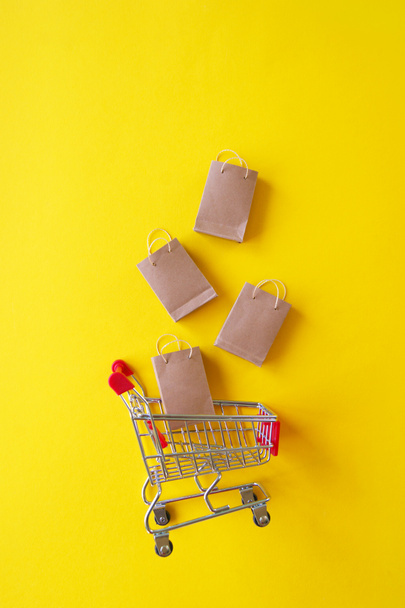 Mini carrinho de compras com pequenos sacos de artesanato no fundo amarelo. O conceito de compras nas lojas e na Internet. - Foto, Imagem