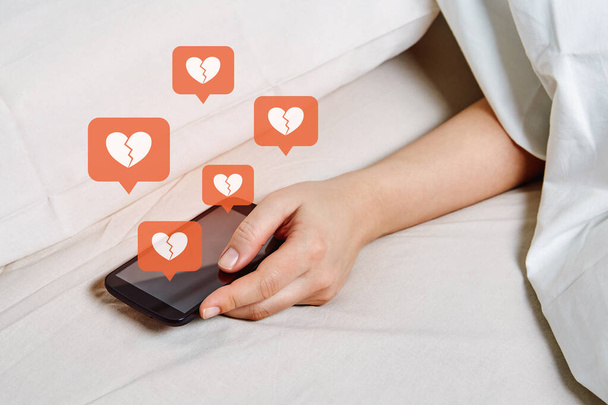 Концепция зависимости и зависимости социальных сетей. Депрессивная девушка под одеялом держит смартфон и чувствует себя непопулярным в социальных сетях. - Фото, изображение