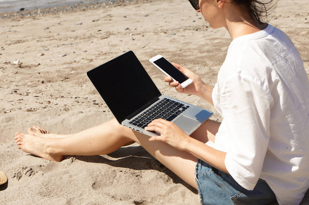 Mujer joven usando un ordenador portátil y sosteniendo un teléfono celular en una playa. Comunicación de estilo de vida vacacional. Concepto de trabajo independiente - Foto, Imagen