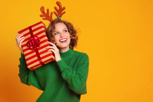 fröhlich komisch überraschte junge Frau in grünem Pullover mit Hirschgeweih hält ein Weihnachtsgeschenk in der Hand und lacht auf gelbem Hintergrund - Foto, Bild