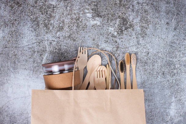 Basic set eco friendly bamboo cutlery, reusable coffee cup, toothbrushes, metal tubes on paper shopping bag, gray background. Concept of environmental protection, plastic free, zero waste. Copy space - Photo, Image